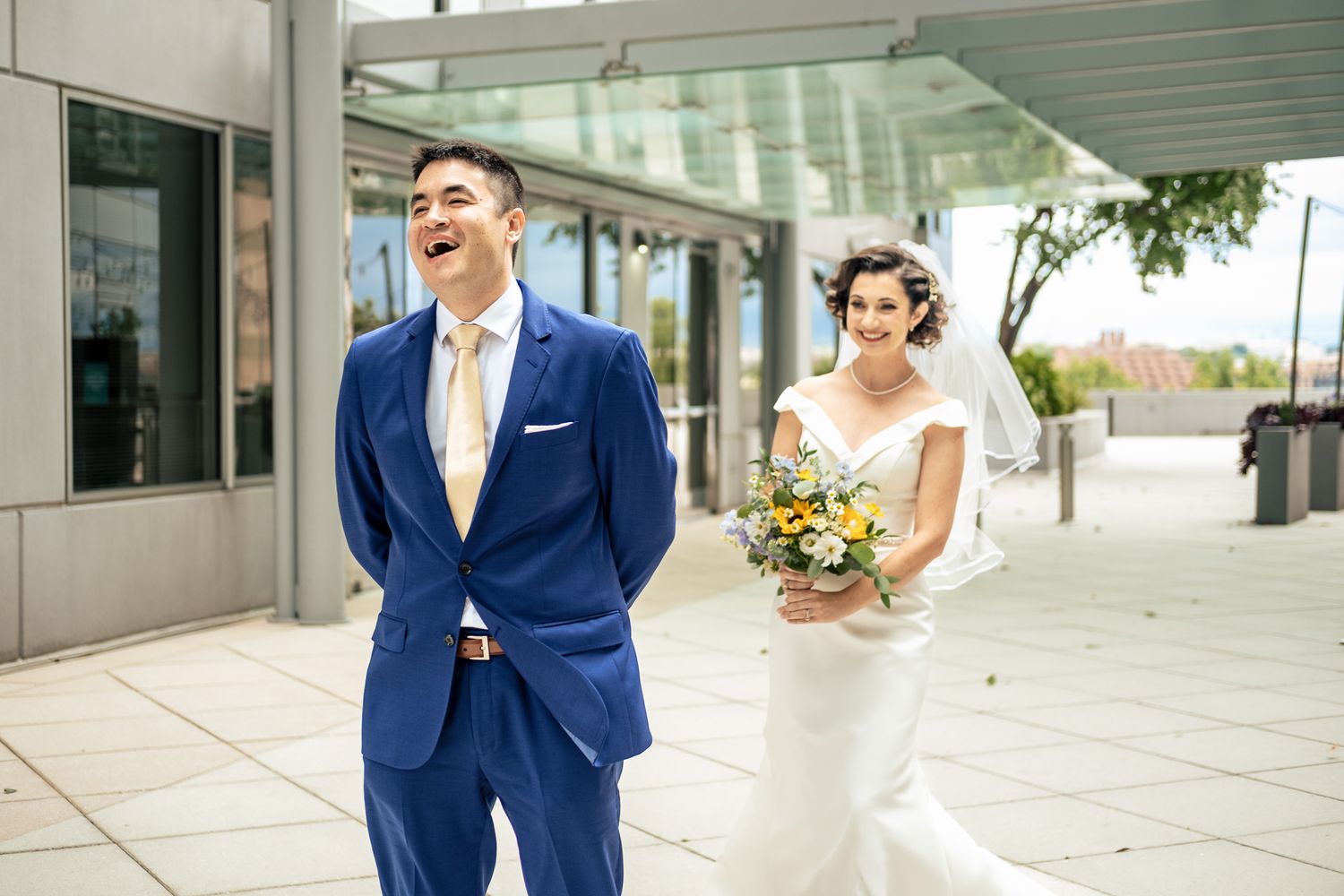 Groom in blue suit laughs joyfully during first look as bride approaches holding yellow bouquet.