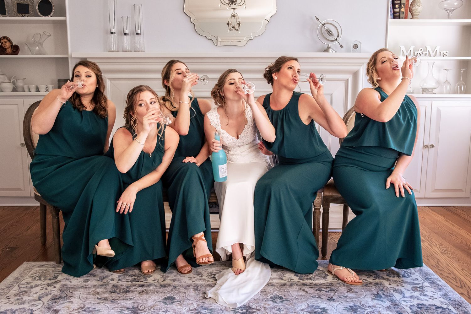 Bride and five bridesmaids in emerald green dresses blow kisses together indoors.