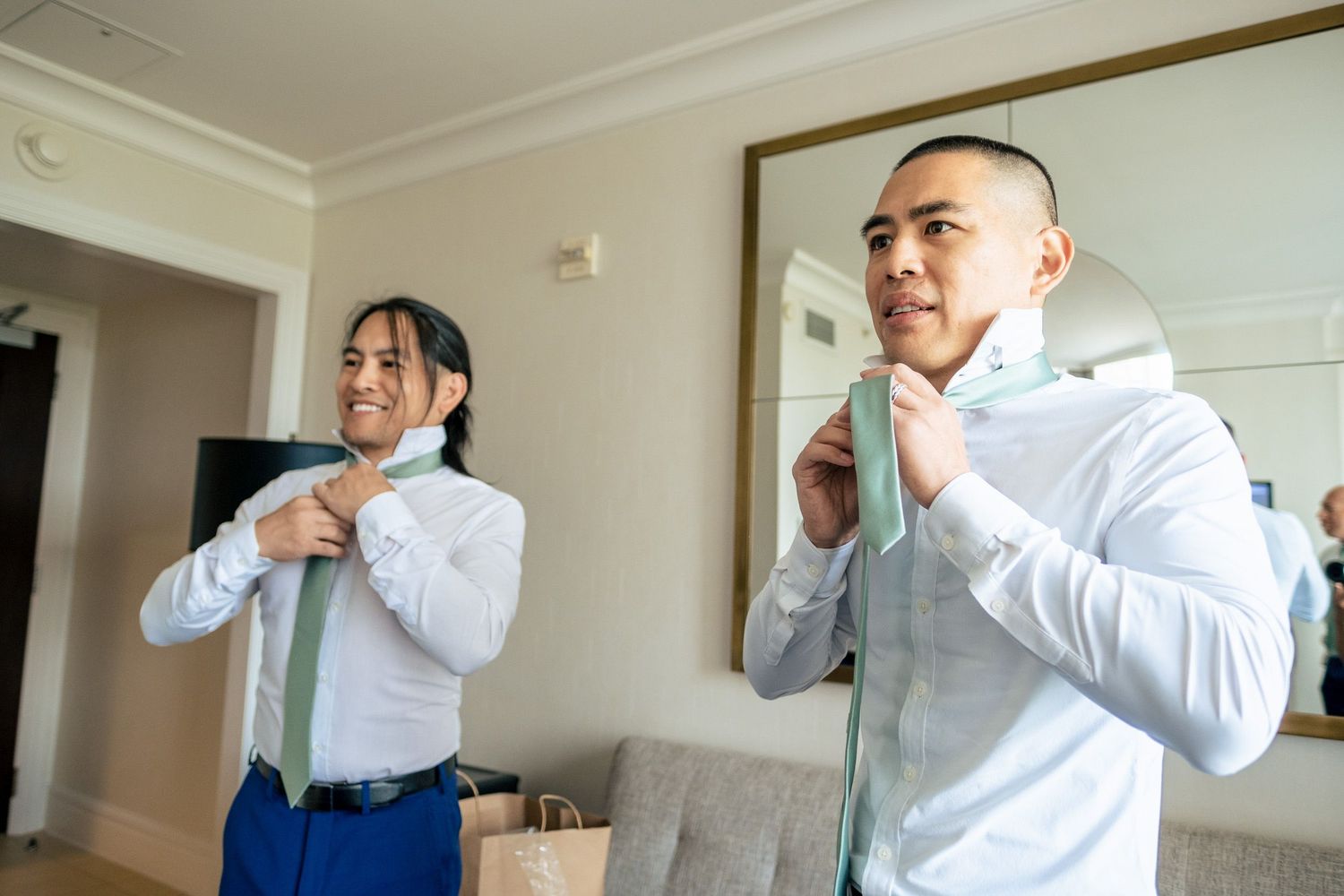 Two groomsmen in blue suits helping each other tie their green ties before a wedding.