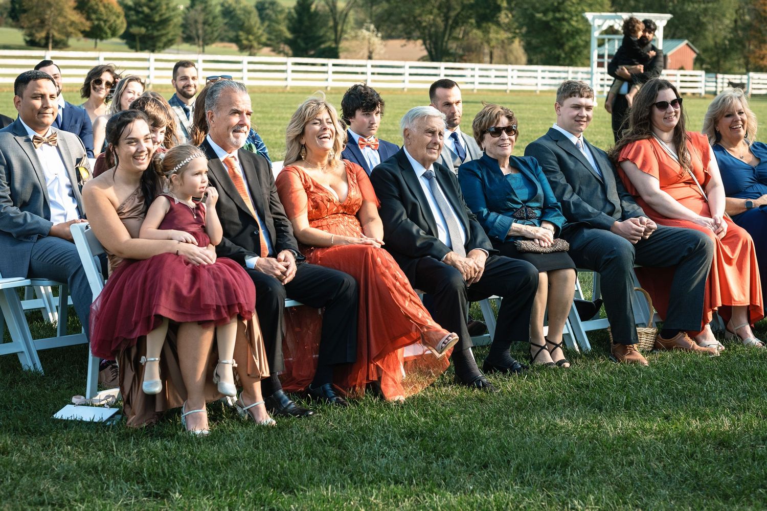 Smiling wedding guests including family members seated in white chairs at an outdoor farm ceremony.