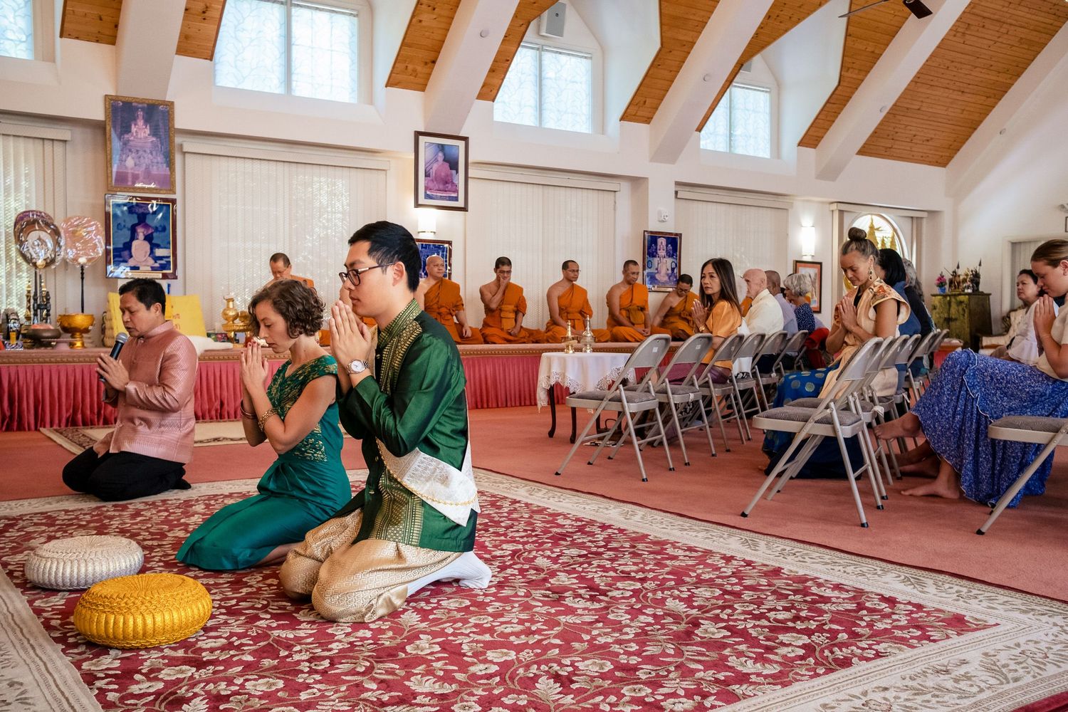 Couple kneels in prayer before Buddhist monks in saffron robes during traditional Thai wedding ceremony.