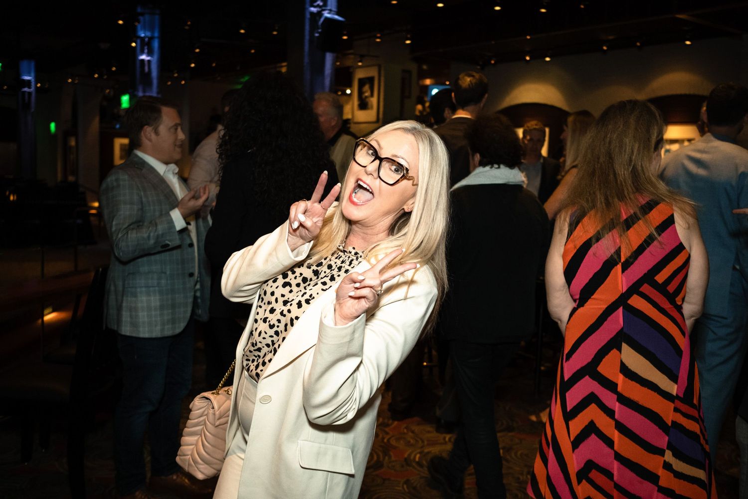 Blonde woman in white blazer making peace signs at a lively indoor party with guests in colorful attire.