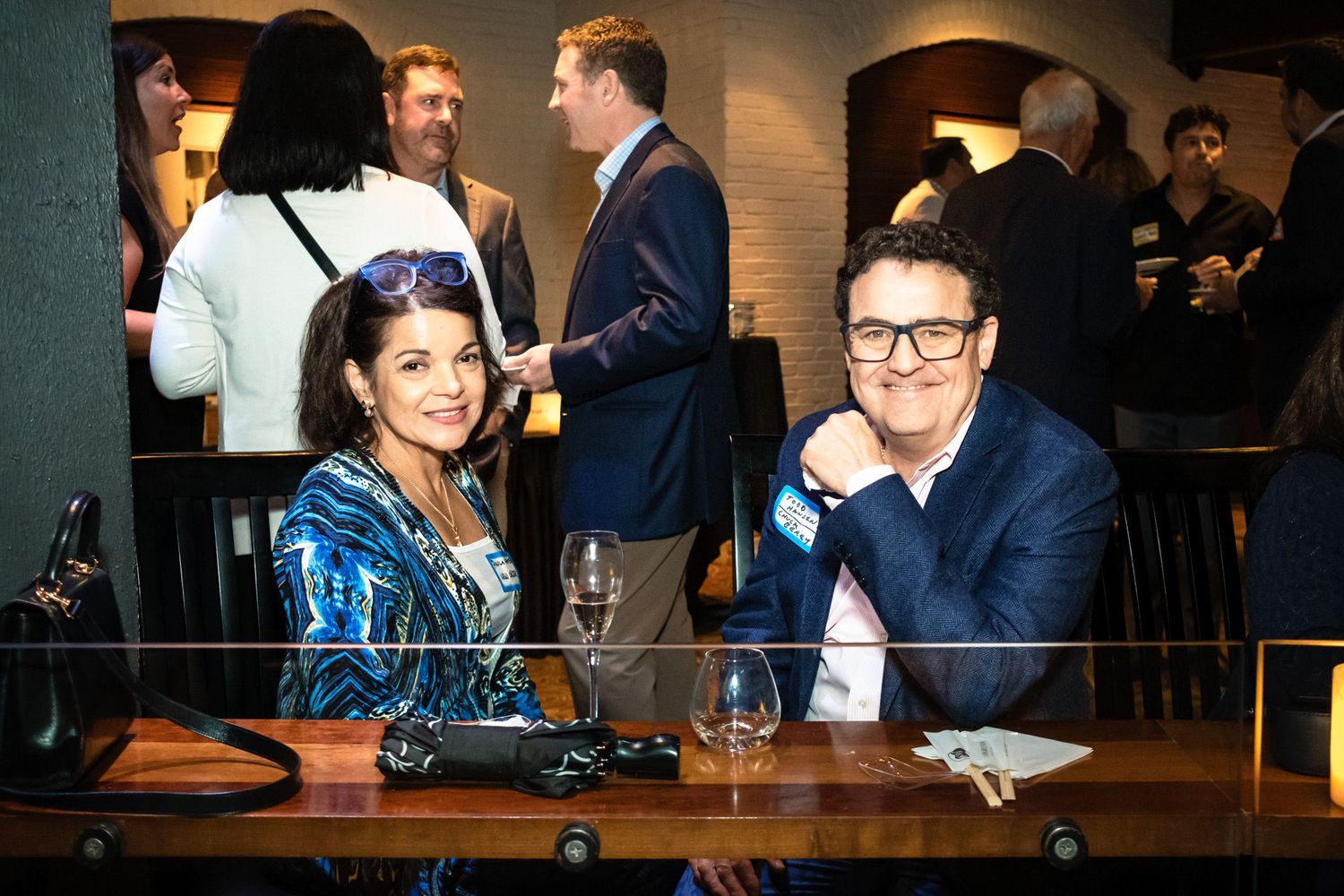 Two smiling guests seated at a bar during an elegant networking event with attendees socializing in the background.