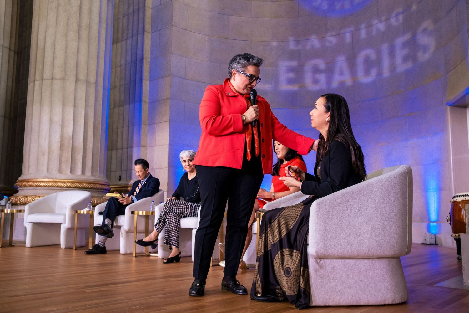 Man in red jacket adjusts microphone for woman at Lasting Legacies event on elegant stage.