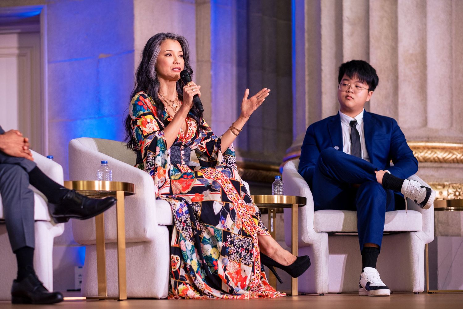 Woman in floral dress speaks on panel at elegant event, seated beside man in blue suit on stage.