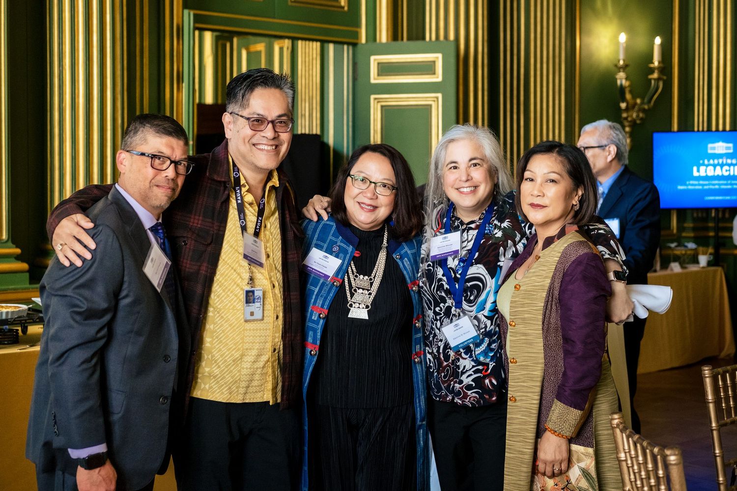 Five professionals smile together at a formal networking event in an elegant green-paneled room.