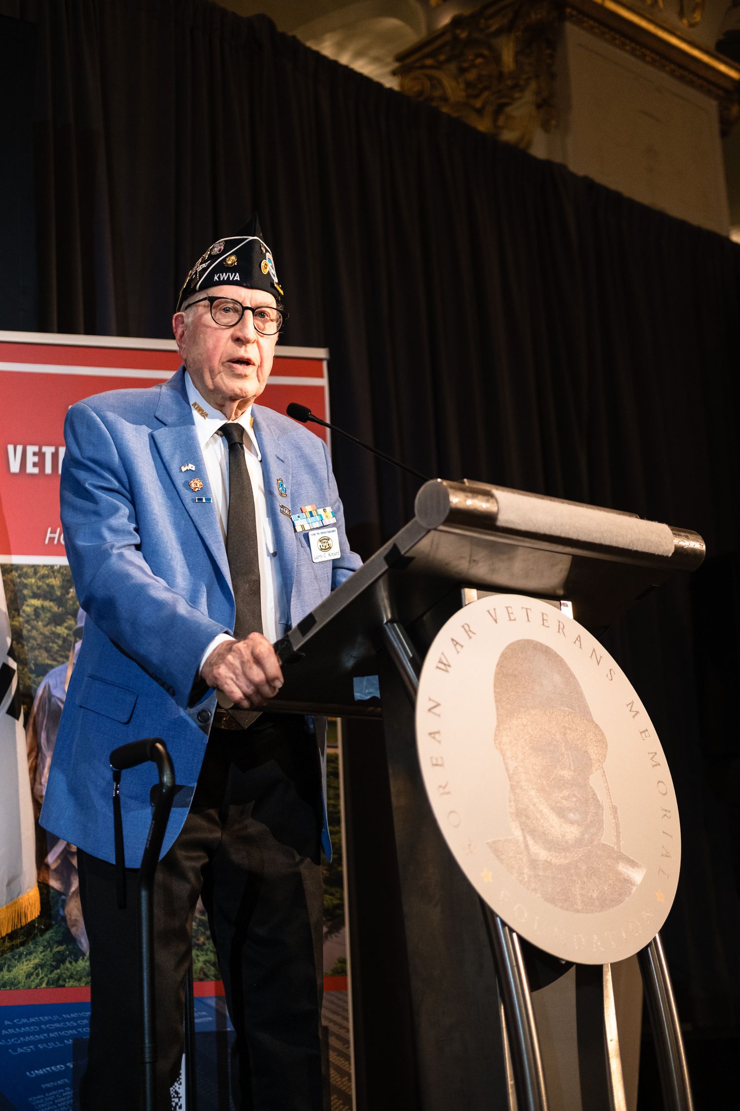 Elderly veteran in blue blazer and military cap speaks at podium at Korean War Veterans Memorial Foundation event.