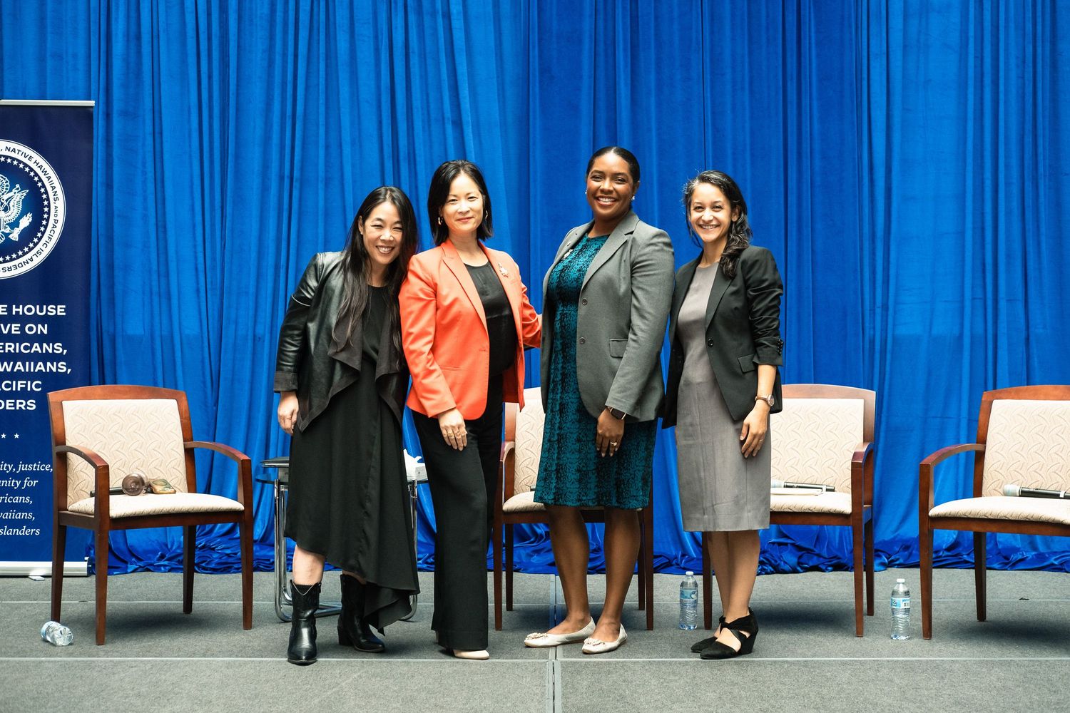 Four professional women pose together on stage in front of a blue curtain at a White House policy event.