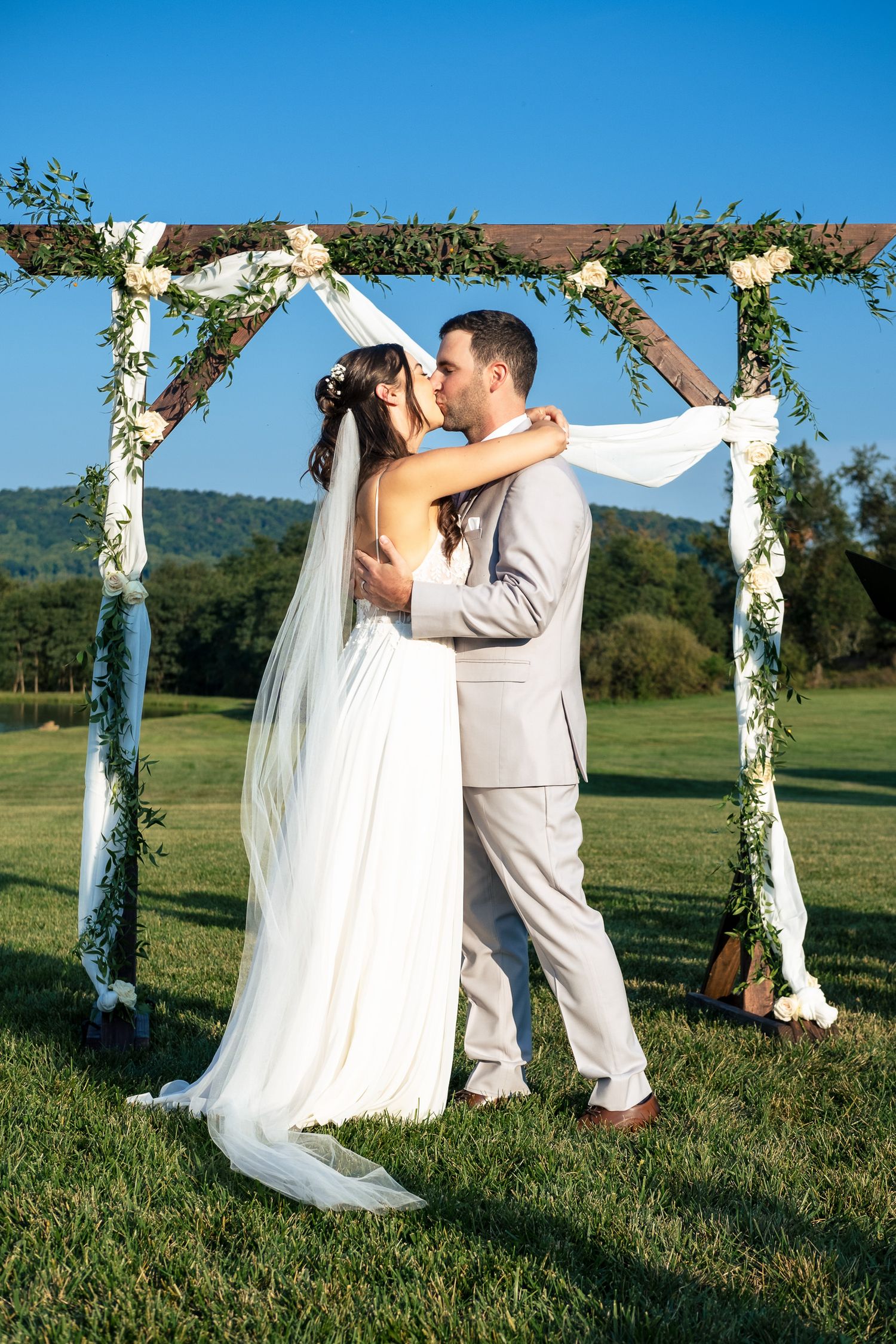 Bride and groom share their first kiss under a rustic wooden arch adorned with white flowers and greenery.