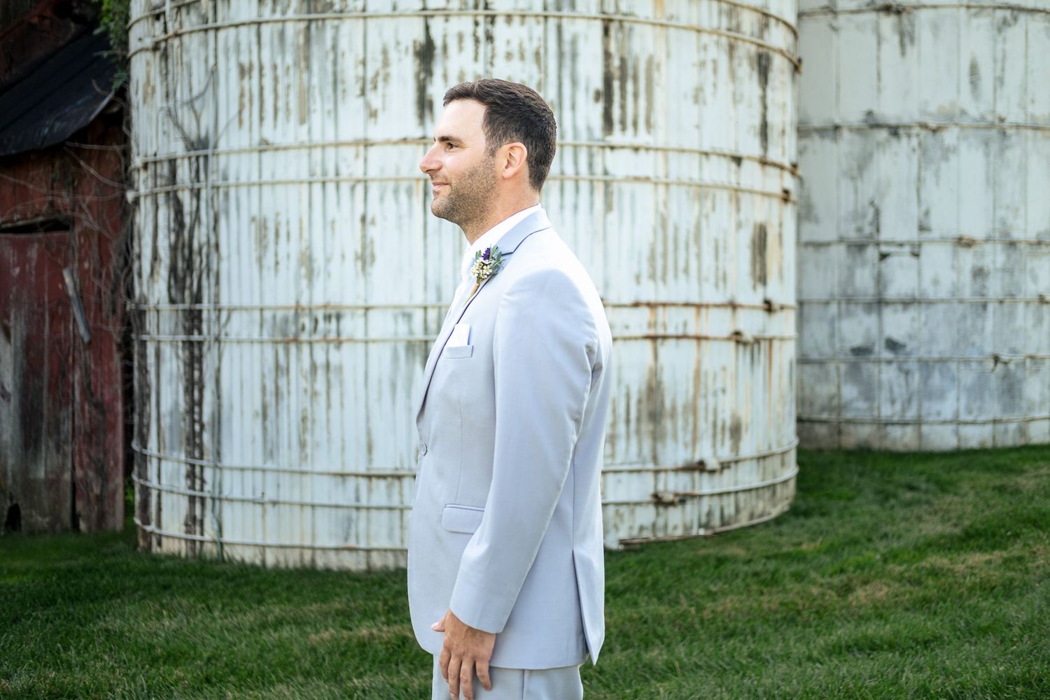 Dapper groom in light gray suit standing profile beside vintage rustic grain silos outdoors.