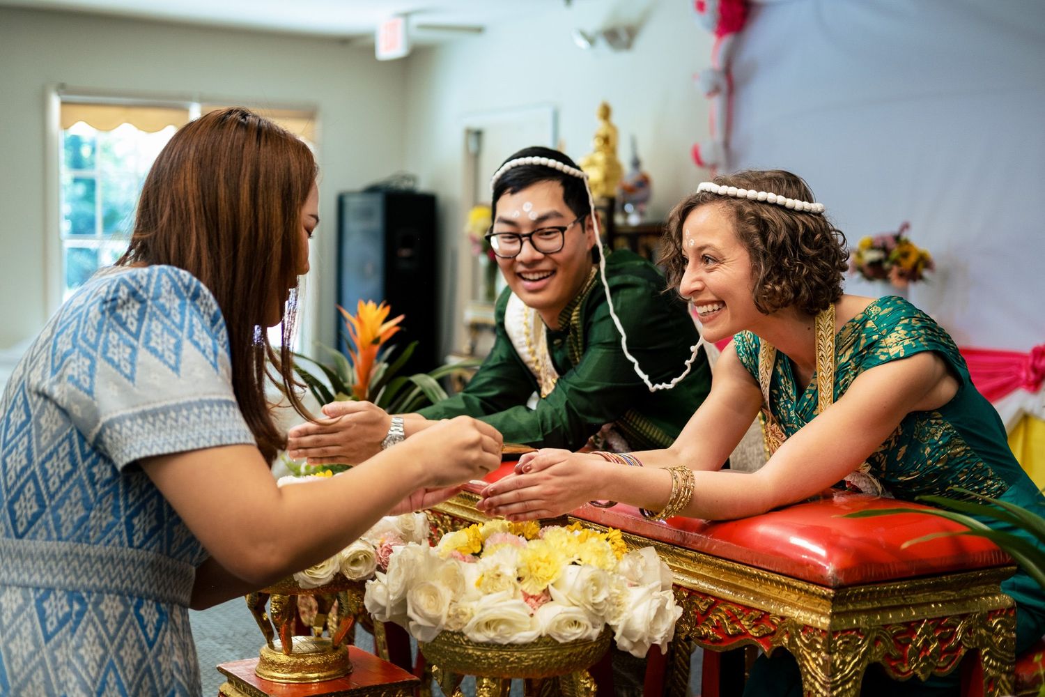 Two guests smiling while presenting flowers to couple at traditional Thai Buddhist wedding ceremony.