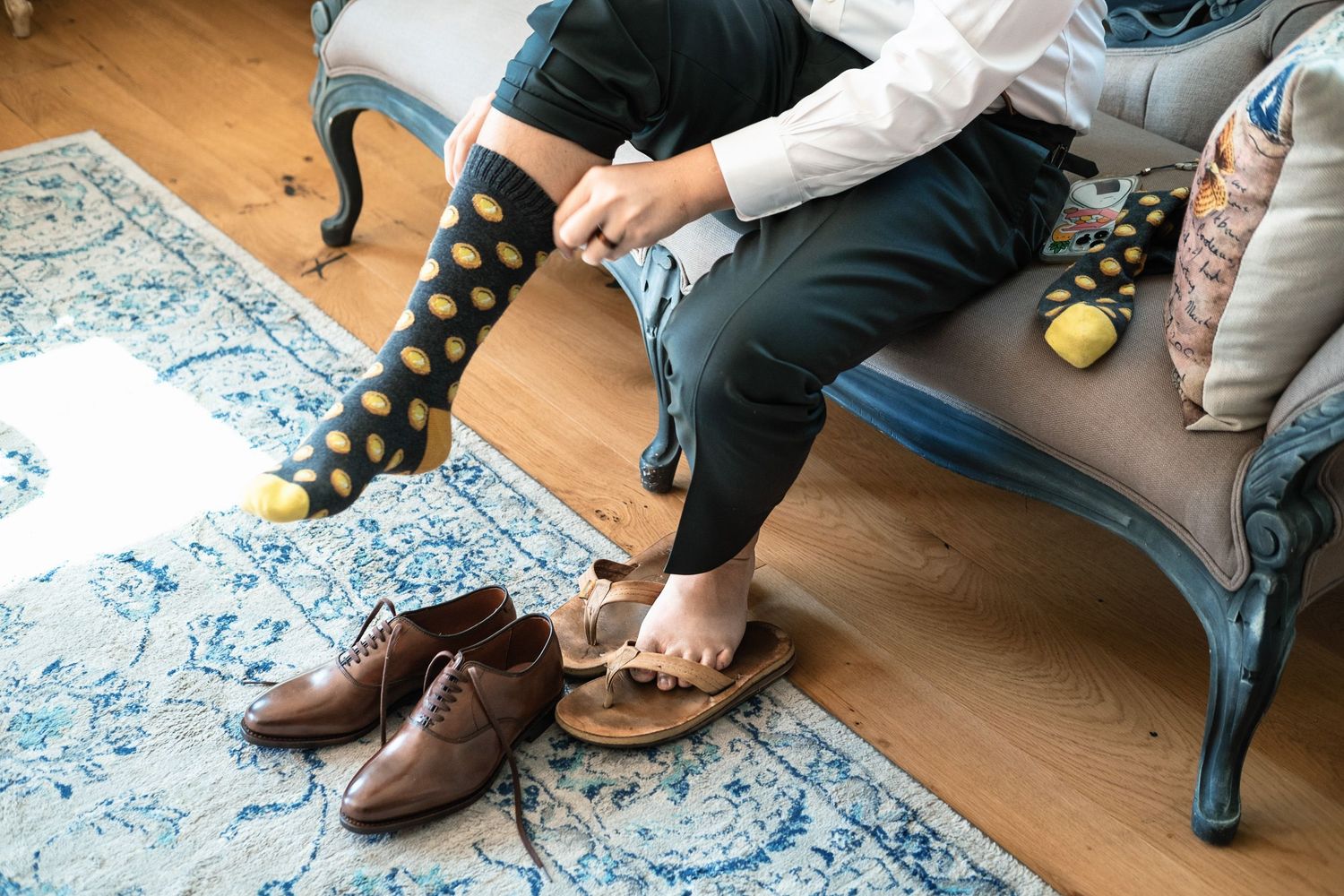 Groom putting on fun polka dot socks beside brown oxford dress shoes on rug.