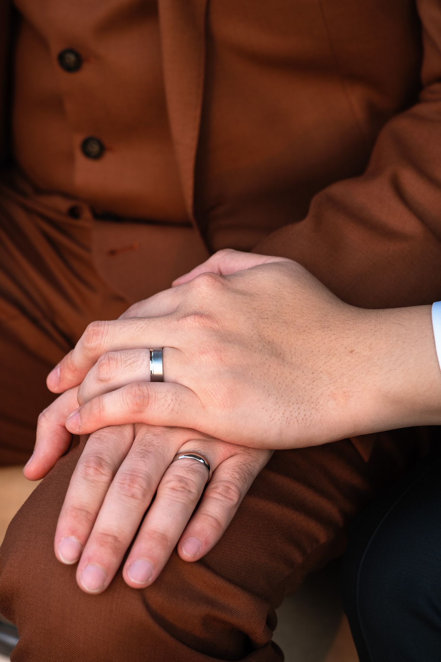 Close-up of two grooms holding hands showing matching wedding bands, one in a brown suit.
