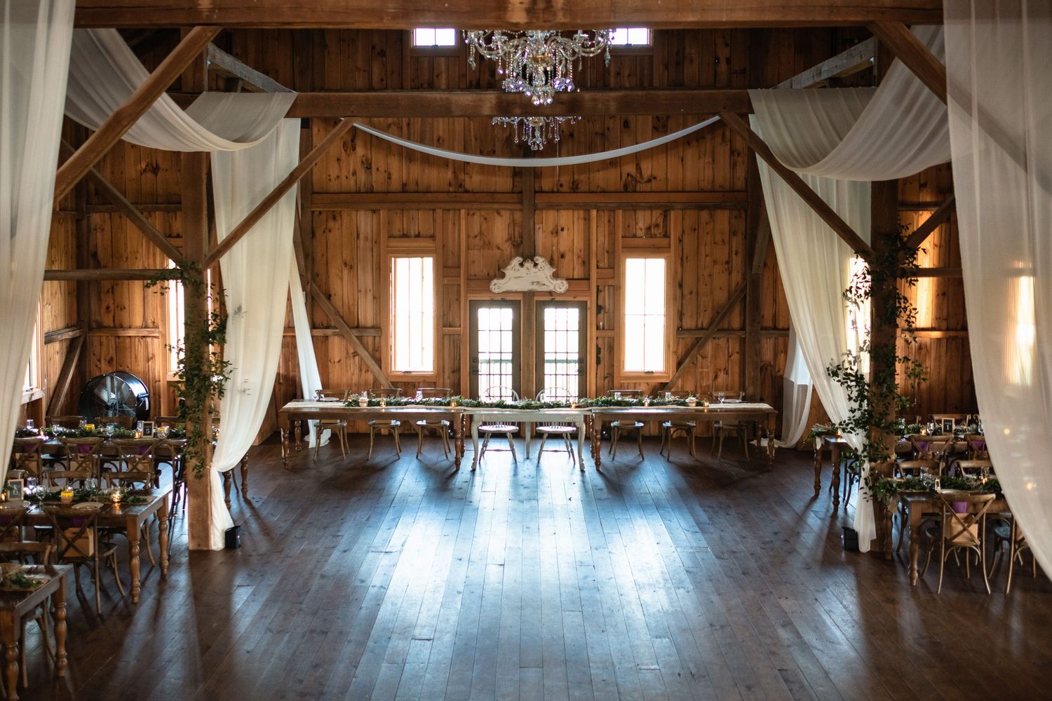 Elegant rustic barn wedding reception with draped white fabric, chandeliers, and decorated dining tables.