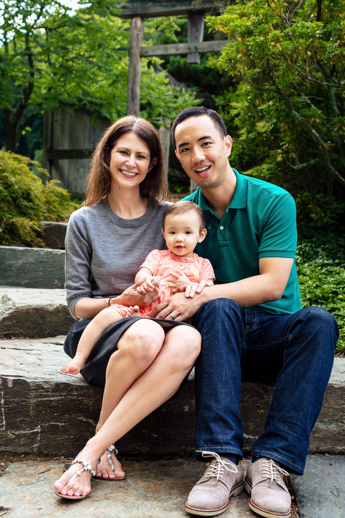 Happy family portrait with parents and baby sitting on stone steps in a lush garden setting.