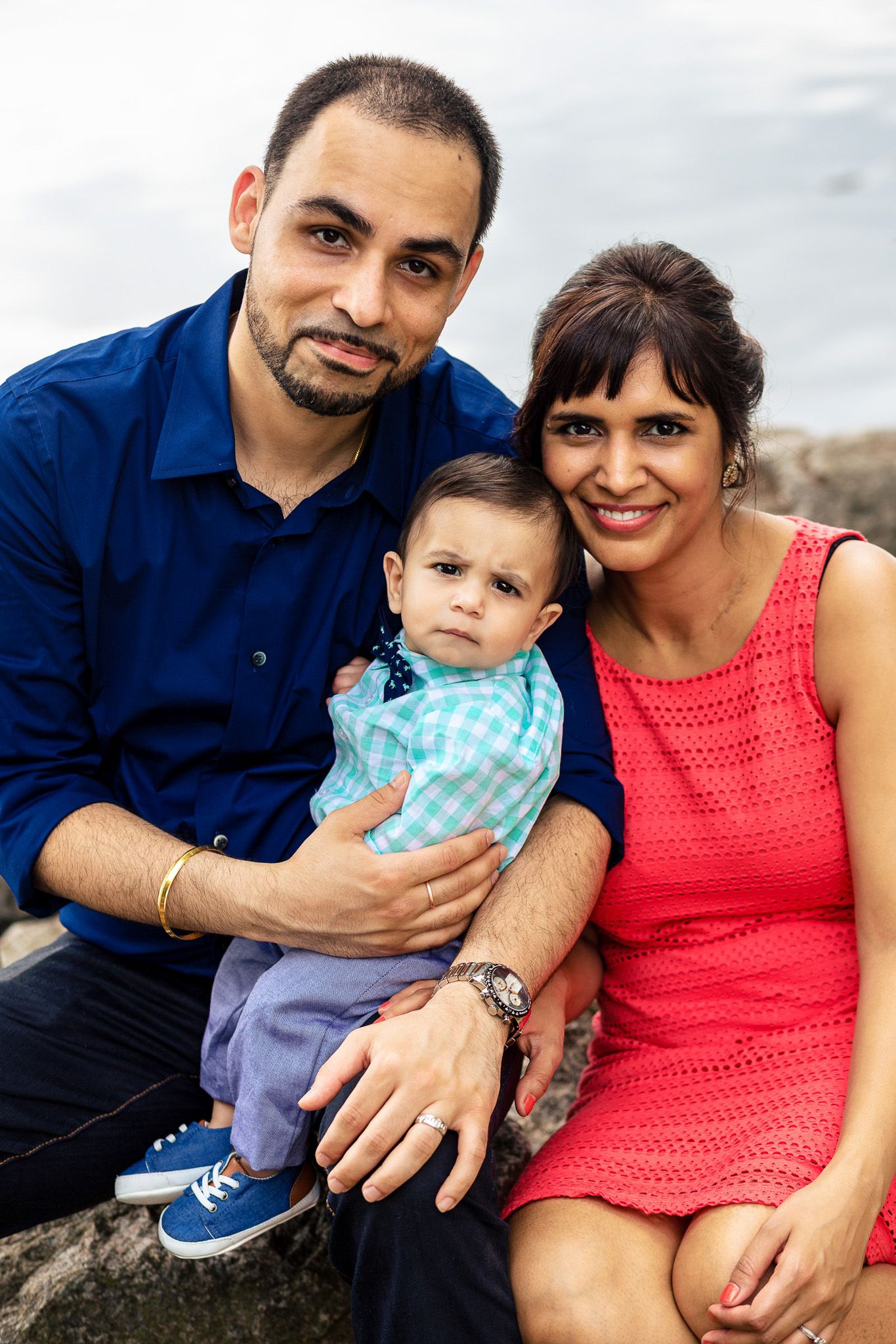 Young family of three posing together outdoors, father and mother holding their baby boy by the water.