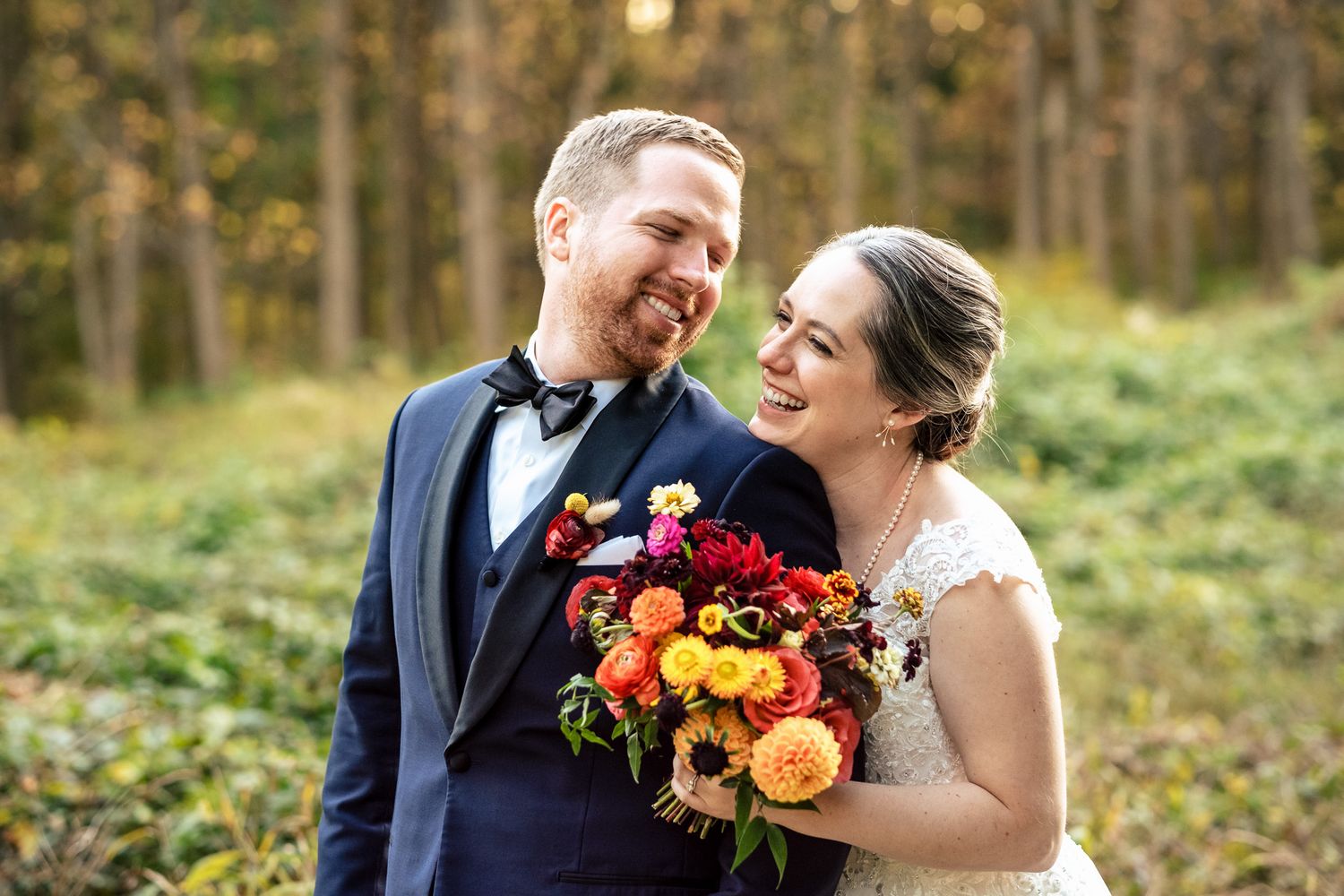 Happy couple embracing in autumn forest, bride holding vibrant red and orange bouquet.