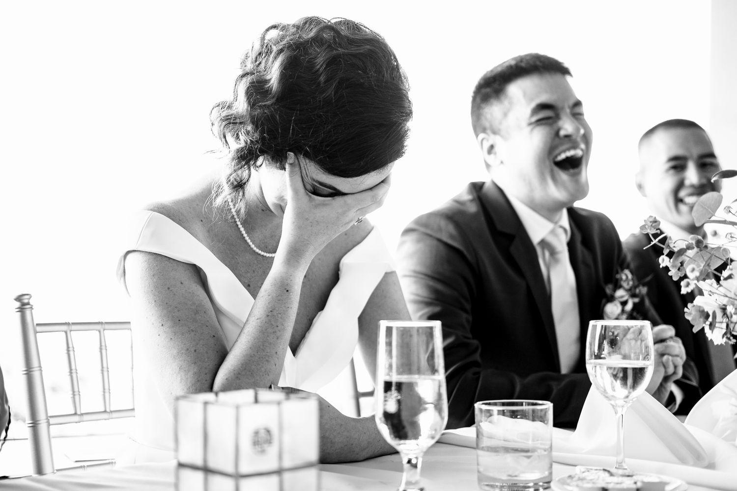 Bride and groom laugh joyfully at their wedding reception table in black and white photo.
