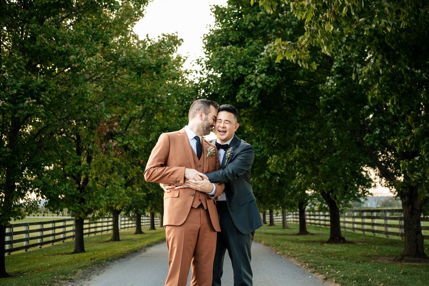 Two grooms in suits sharing a tender moment on a tree-lined path at golden hour.