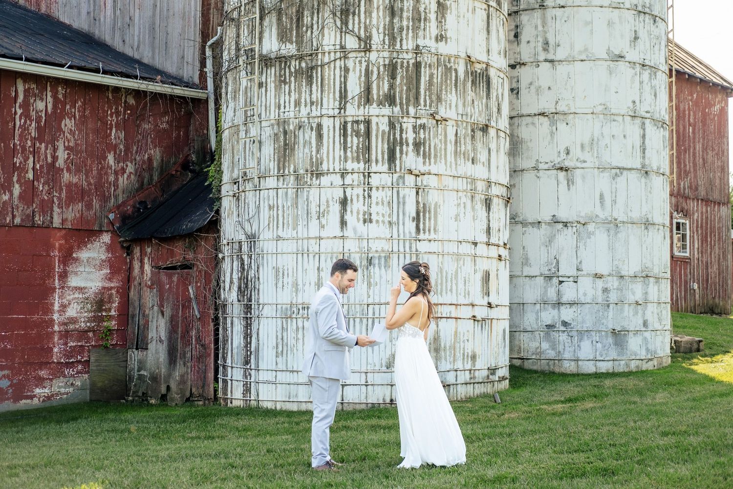 Bride and groom hold hands smiling at each other beside rustic red barn and grain silos.