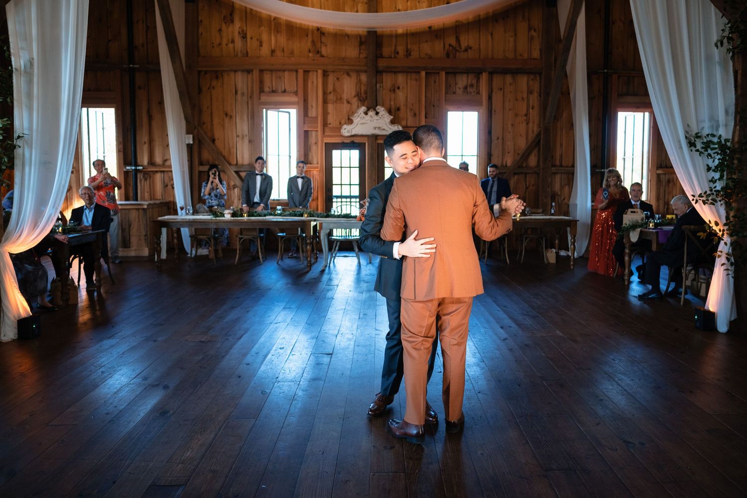 Two grooms share first dance in rustic barn wedding venue with guests watching.
