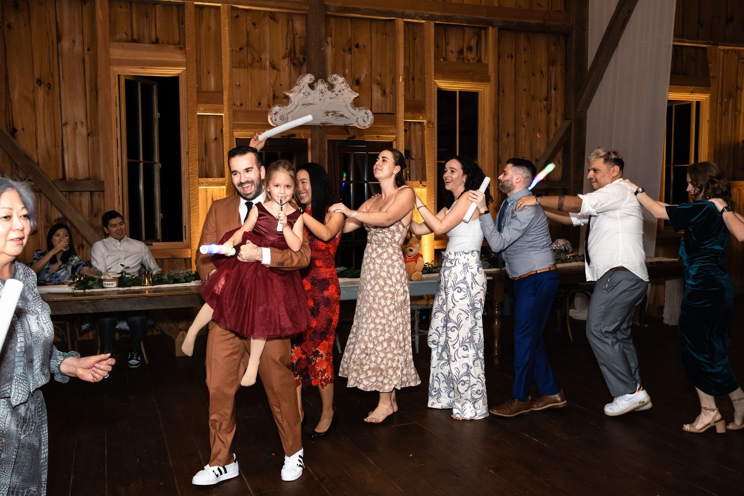 Guests and groom in brown suit dance together in a line inside a rustic barn reception.