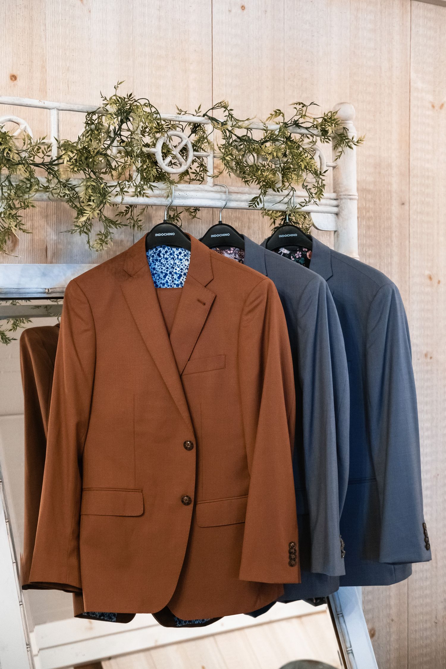 Brown, charcoal, and gray wedding suits hang on a rack decorated with greenery in a rustic venue.