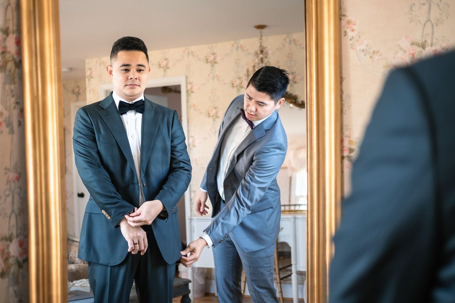 Groomsman helping groom button navy tuxedo jacket while checking mirror reflection.