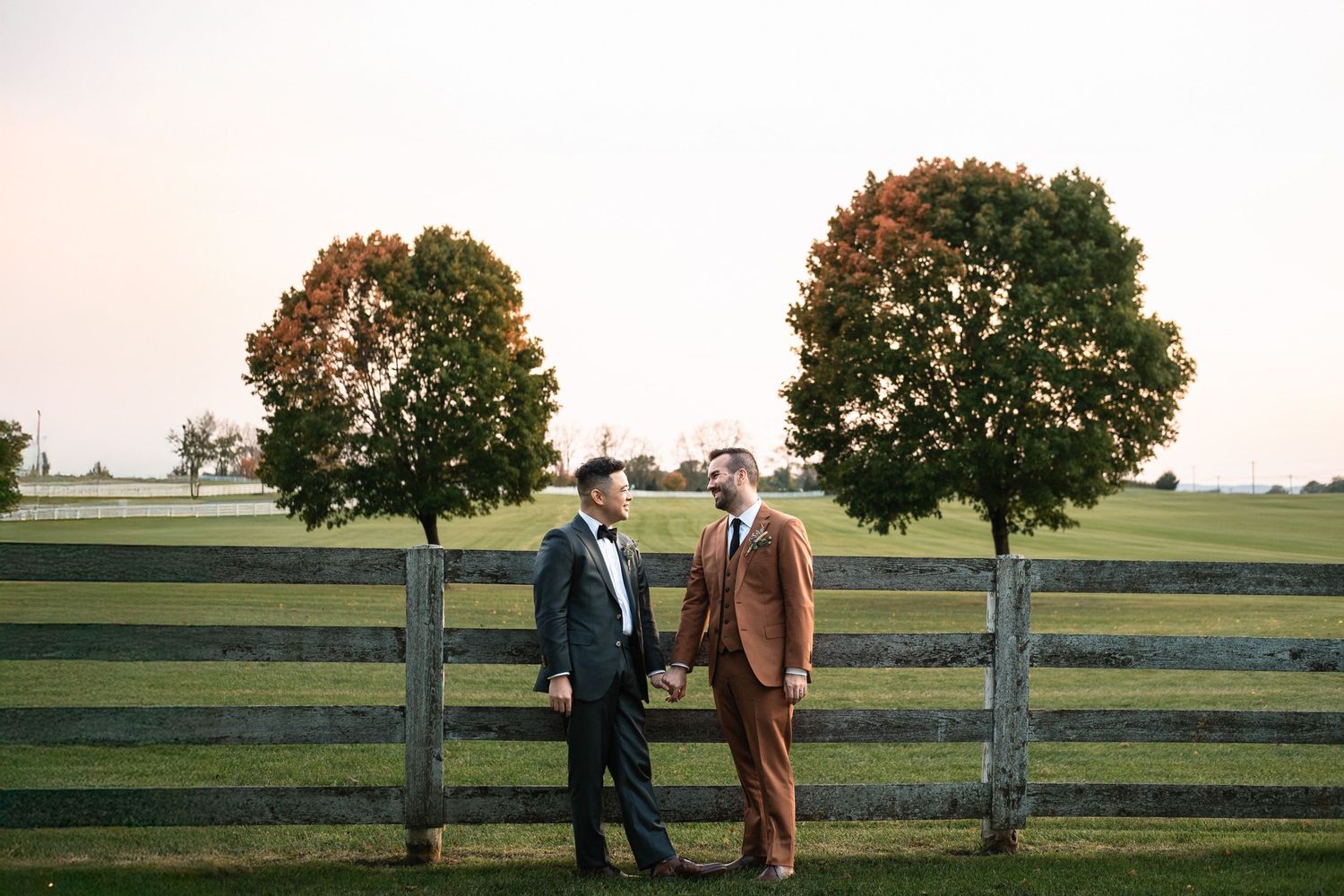 Two grooms hold hands near a wooden fence with autumn trees at golden hour on their wedding day.