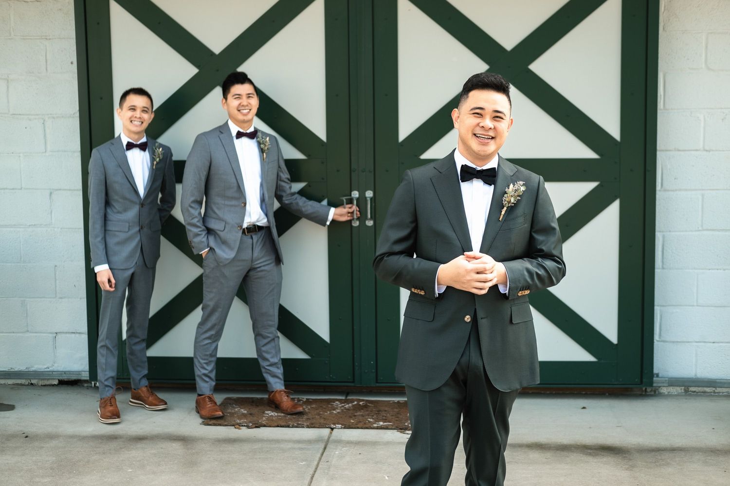 Three groomsmen in gray suits and black bow ties posing outside barn wedding venue doors.