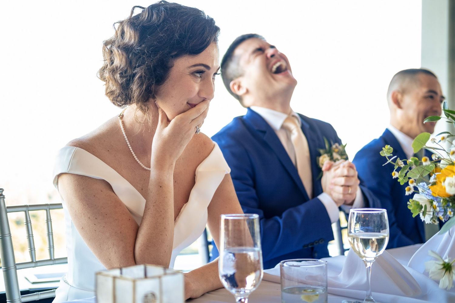 Bride rests her chin on her hand while groom laughs joyfully during wedding speeches at the reception.