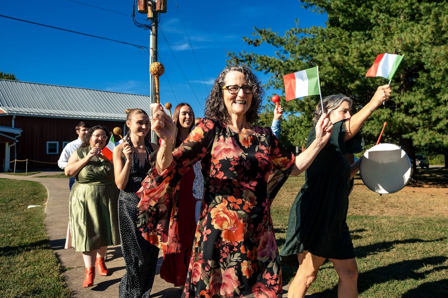 A woman in a floral dress leads a cheerful procession of guests waving Italian flags at a wedding.