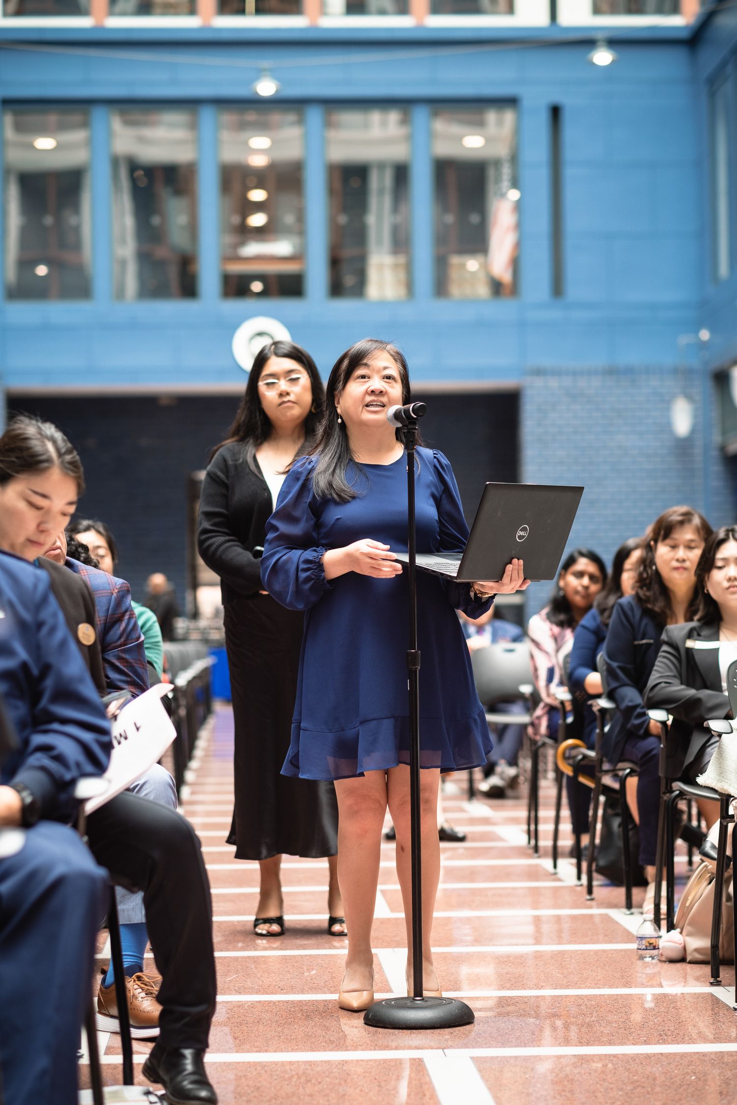 Woman in blue dress speaking at microphone during formal indoor conference event.