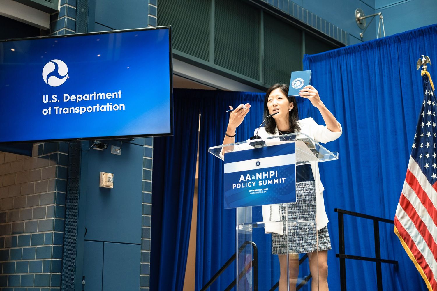 Woman speaks at U.S. Department of Transportation podium during AA+NHPI Policy Summit holding a blue book.