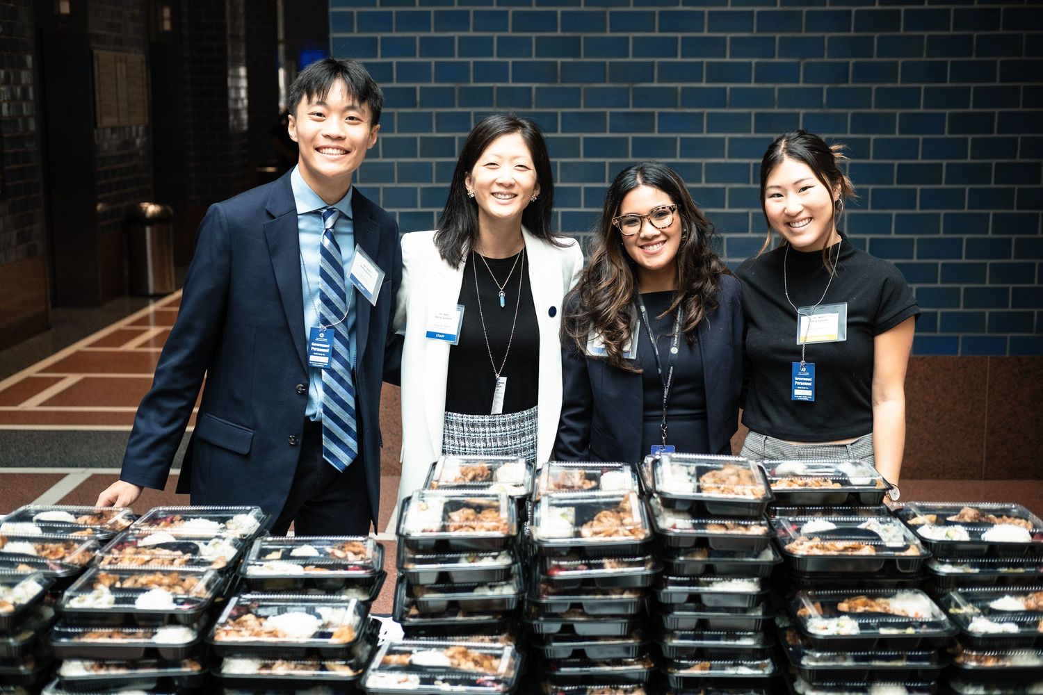 Four professionals smiling behind rows of bento box meals at a catered corporate or charity event.