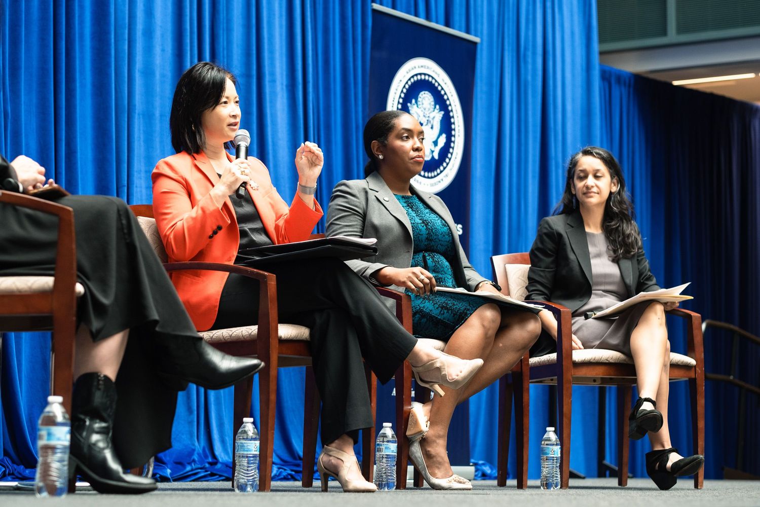 Three professional women seated on stage at a formal panel discussion in front of a blue presidential backdrop.