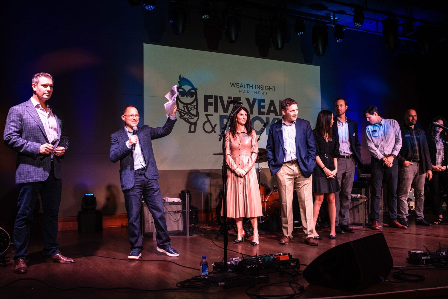 Group of professionals on stage at a Five Year Anniversary awards ceremony event under colorful stage lighting.