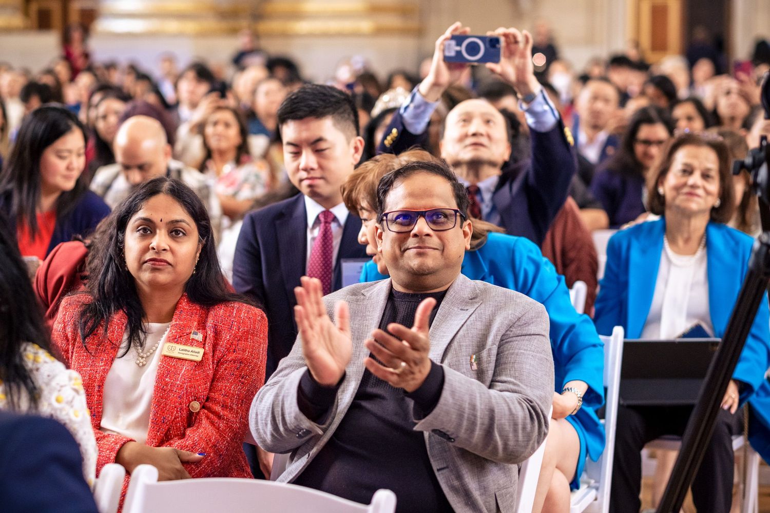 Diverse audience applauding at a formal event, man in gray blazer clapping, woman in red dress seated beside him.