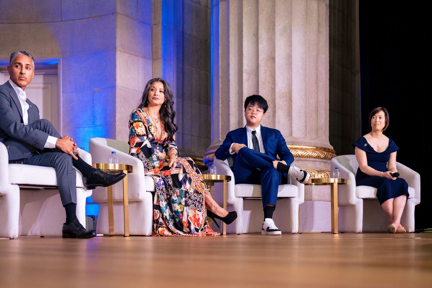 Four panelists seated on stage at an elegant event with blue-lit columns in the background.