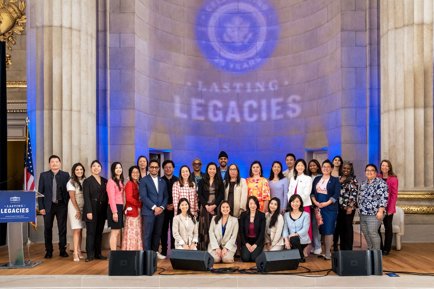 Group photo at Lasting Legacies event in grand hall with blue lighting and official seal backdrop.