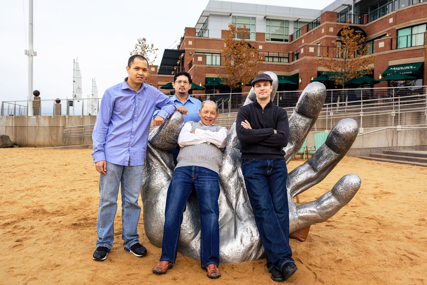 Four men posing with a large metal hand sculpture in an urban outdoor plaza.