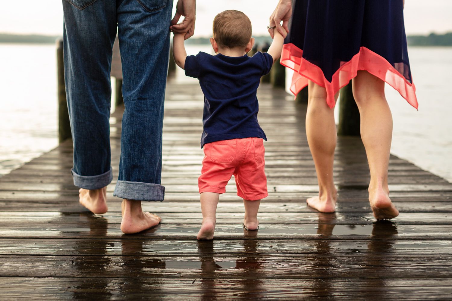 Young toddler walking hand-in-hand with parents on a wet wooden dock overlooking a calm lake.