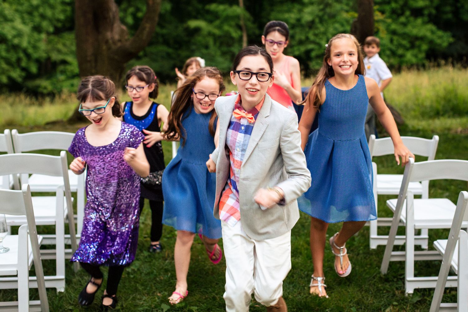 Children running joyfully through outdoor wedding ceremony chairs on green lawn.