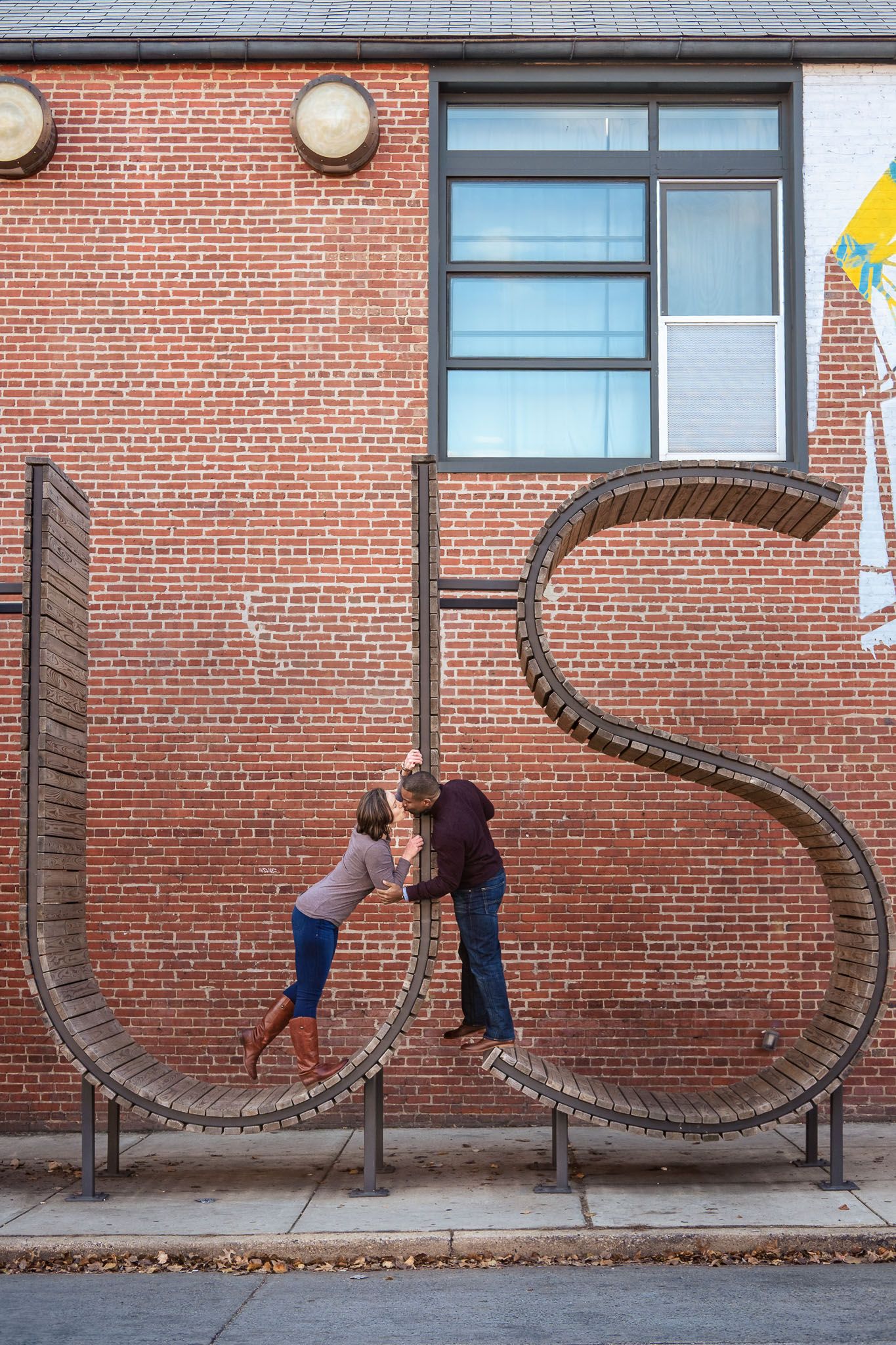 Couple kisses playfully inside large metal US letter sculpture on brick wall.