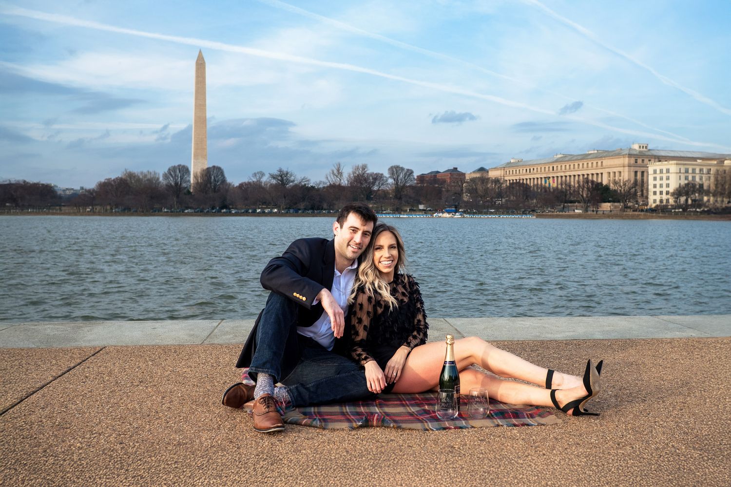Smiling couple sitting on a blanket by the Tidal Basin with the Washington Monument at sunset.