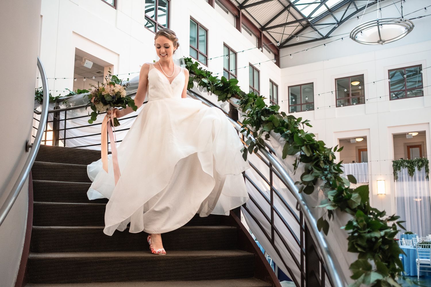 Elegant bride in flowing white gown descends staircase decorated with lush green garland.