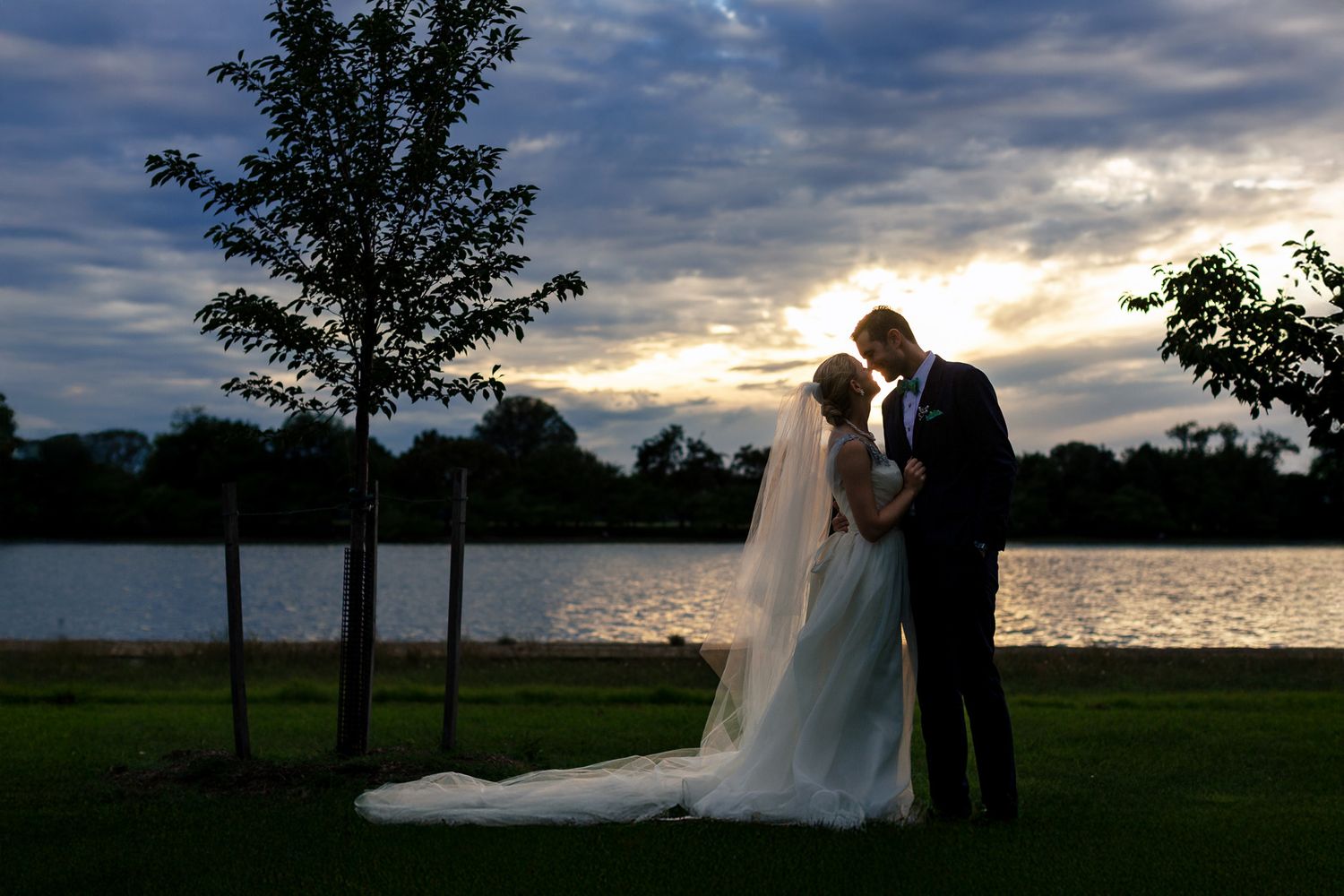 Bride and groom share an intimate moment by a lake at sunset with dramatic cloudy sky.