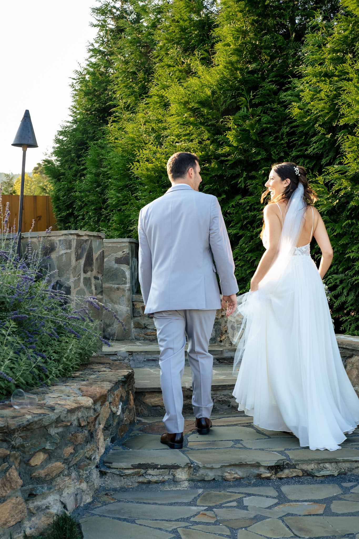 Bride and groom walking hand in hand up stone steps surrounded by lush greenery.