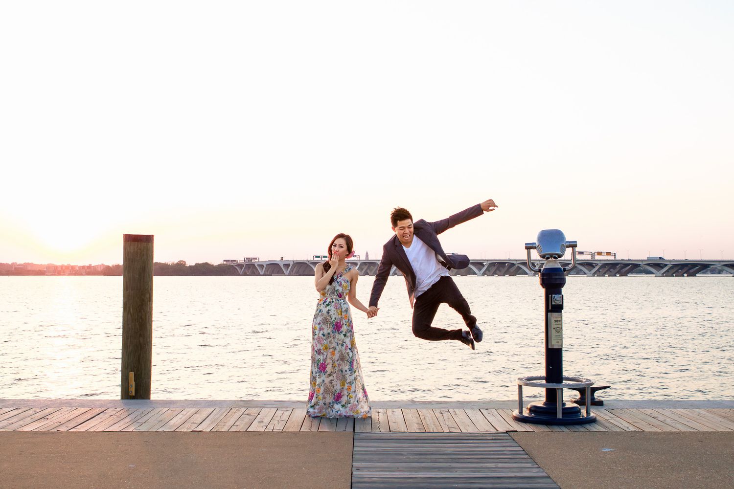Joyful couple holding hands on a waterfront dock at sunset, man jumping playfully in the air.