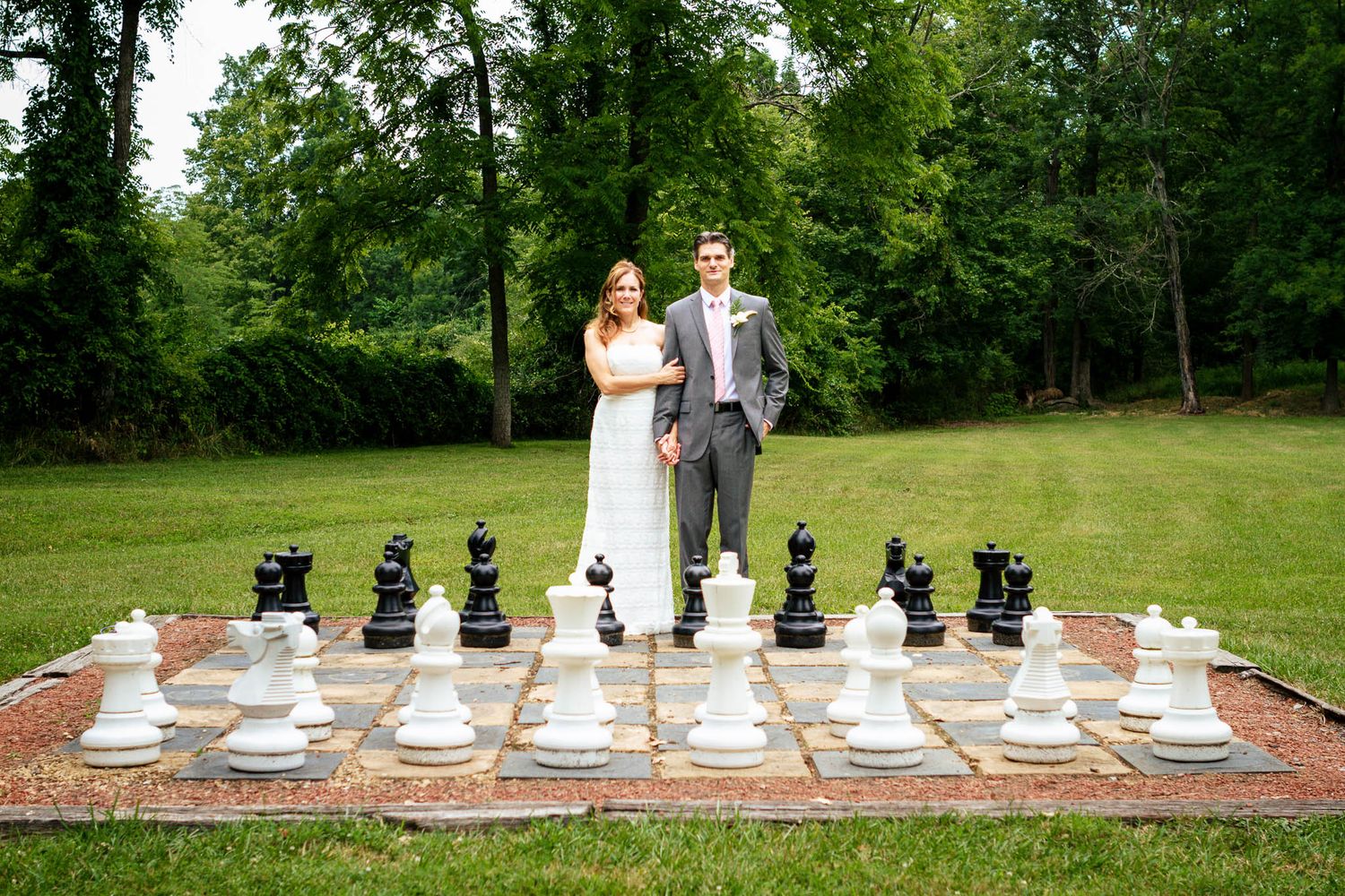 Bride and groom pose together on a giant outdoor chess board surrounded by lush green trees.